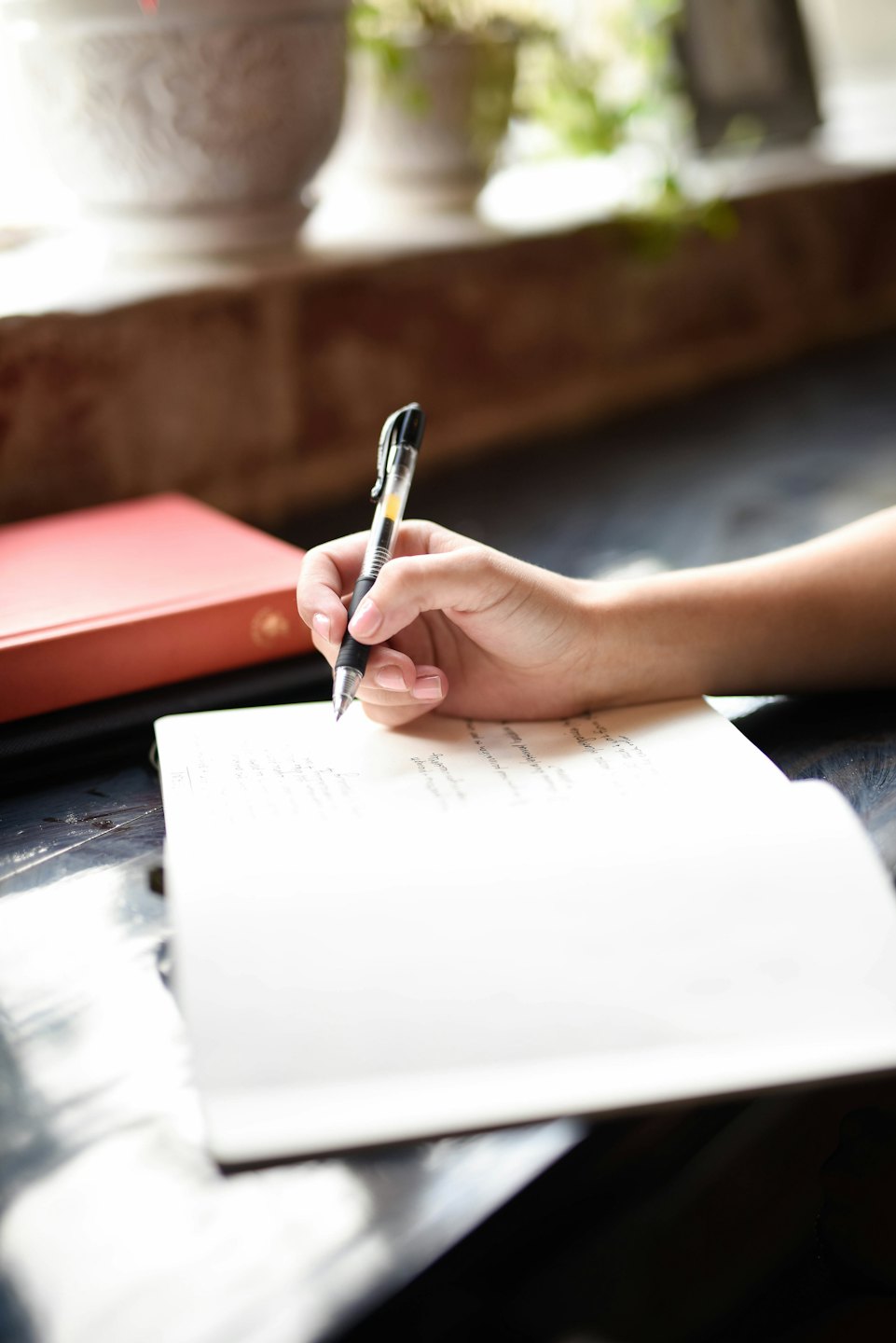 Hand schreibt in Notizbuch mit Stift und rotem Buch.